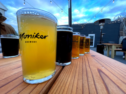 A row of various beers in glasses on a wooden table, with string lights and a cloudy sky in the background.