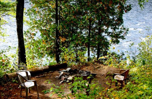 A serene campsite by a river, featuring two chairs around a fire pit surrounded by trees and autumn foliage.