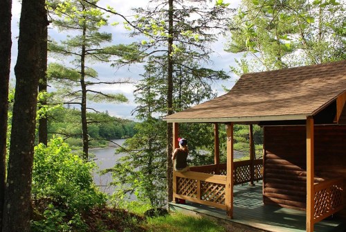 A person sits on the porch of a cabin, surrounded by trees, overlooking a serene lake and cloudy sky.