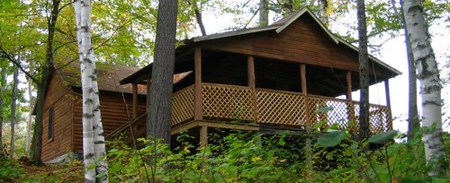 A wooden cabin nestled among trees, featuring a porch with latticework and surrounded by lush greenery.