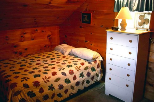 Cozy bedroom with a wooden wall, a bed with a floral quilt, and a white dresser beside a lamp and fan.