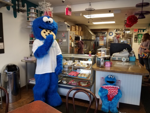 A blue mascot resembling Cookie Monster stands in a bakery, holding a cookie, with a smaller Cookie Monster figure nearby.