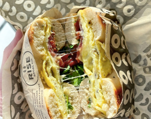 A close-up of a bagel sandwich filled with eggs, bacon, cheese, and greens, wrapped in branded paper.