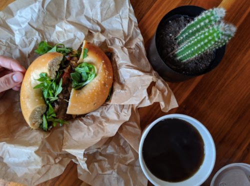 A hand holds a sandwich with greens, next to a cactus in a pot and a cup of coffee on a wooden table.