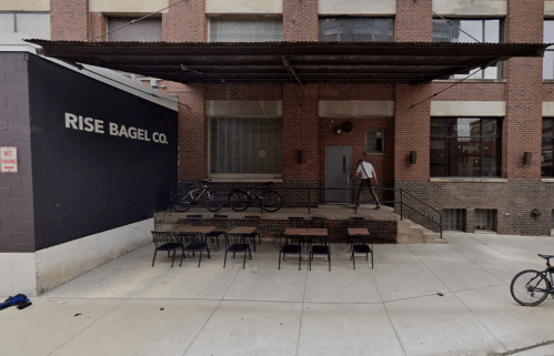 Exterior of Rise Bagel Co. with outdoor seating and bicycles parked nearby. A person is walking up the steps.