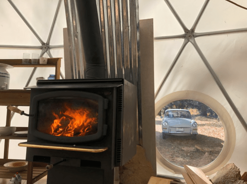 A cozy wood stove inside a geodesic dome, with a vintage car visible through a round window outside.