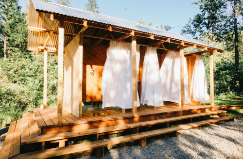A modern wooden cabin with white curtains, surrounded by trees and a gravel path.