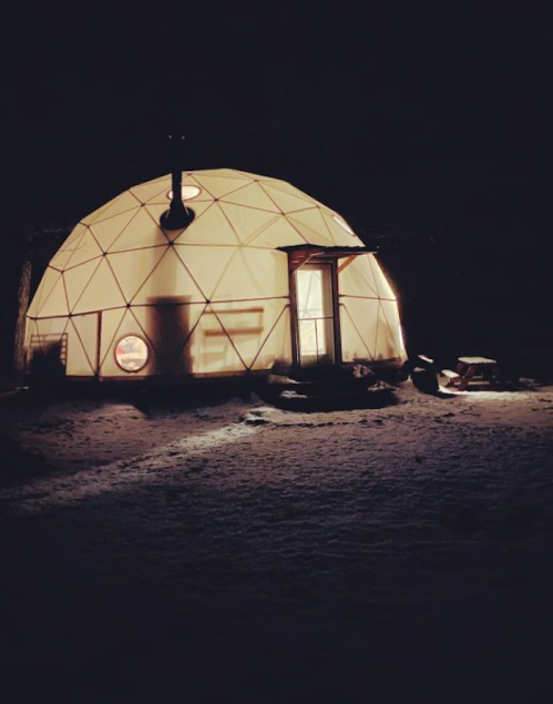 A lit geodesic dome structure at night, surrounded by snow, with a small porch and a warm glow from the windows.