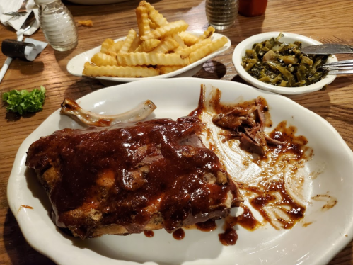A plate with BBQ ribs covered in sauce, a side of crinkle-cut fries, and a dish of greens on a wooden table.