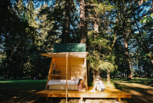 A cozy wooden cabin in a forest, with a child sitting on the deck and an adult nearby, surrounded by tall trees.