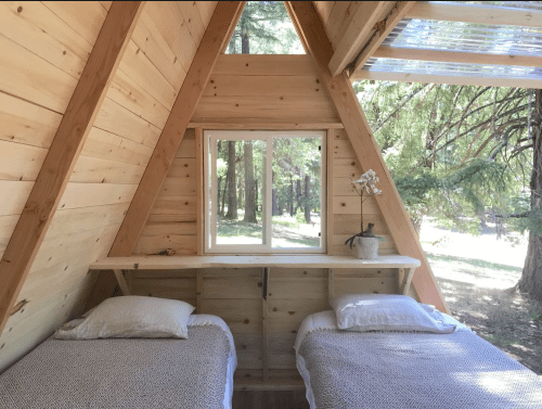 Cozy A-frame cabin interior with two beds, a wooden shelf, and a window overlooking a forested area.