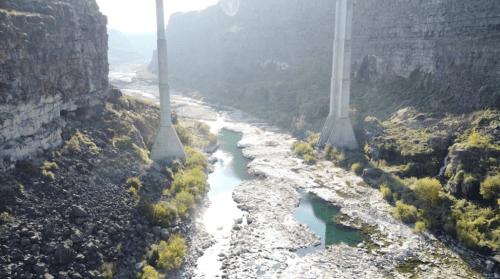 A serene river flows between rocky cliffs, with tall concrete pillars rising above the landscape under a bright sky.