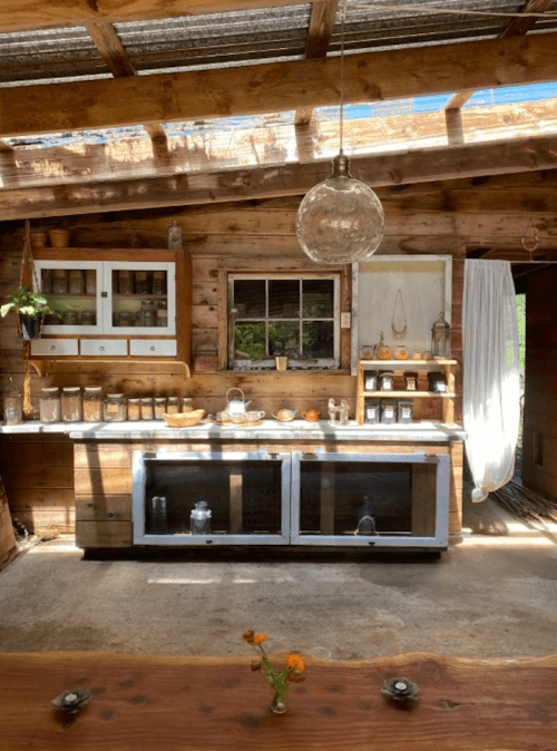 Cozy rustic kitchen with wooden shelves, jars, and a pendant light, featuring a window with greenery outside.