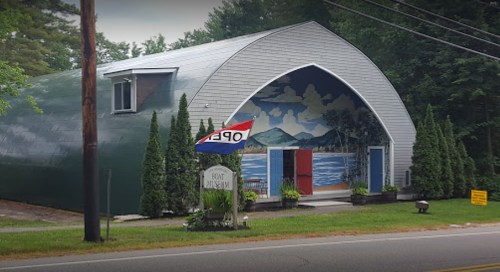 A large, modern building with a curved roof, featuring a mural and an "OPEN" sign in front, surrounded by greenery.