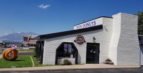 A donut shop with a sign reading "MTN DONUTS," featuring a large donut sculpture outside and mountains in the background.