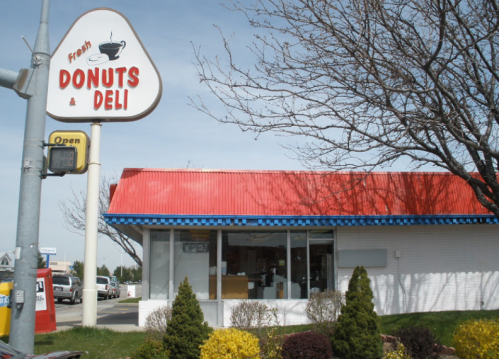 A small deli with a red roof and a sign reading "Fresh Donuts & Deli," surrounded by shrubs and trees.