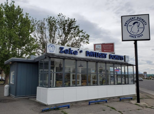 Exterior of Zeke's Daylight Donuts shop, featuring a sign and large windows, with trees and a cloudy sky in the background.