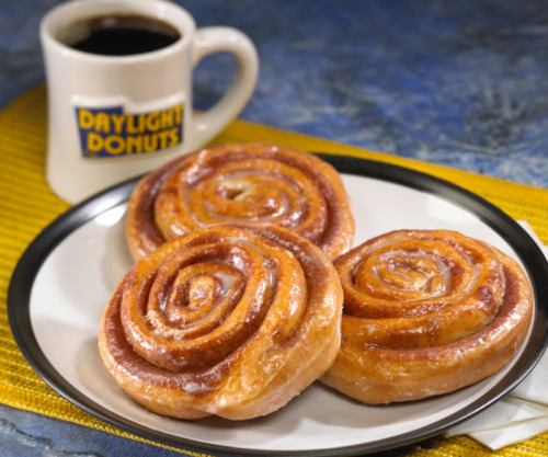 Three glazed cinnamon rolls on a plate next to a coffee mug labeled "DAYLIGHT DONUTS."