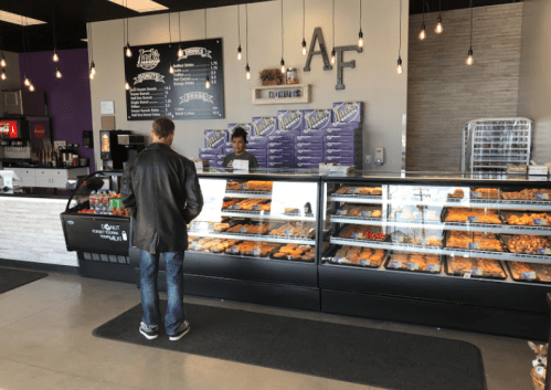 A customer stands at the counter of a donut shop, looking at a display case filled with various donuts.