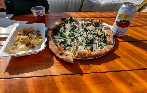 A wooden table with a pizza topped with greens and a side of fried dumplings, plus a can of beer.