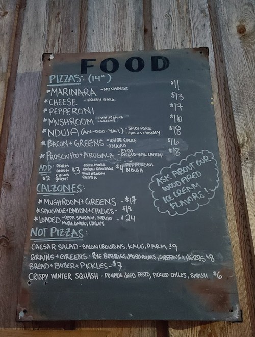 Menu board listing pizzas, add-ons, and non-pizza items with prices, displayed on a rustic background.