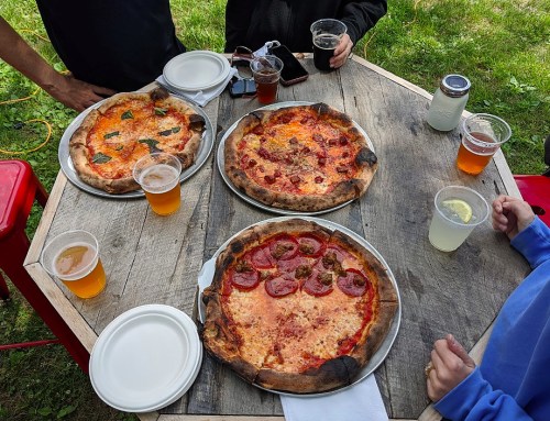 Three pizzas on a wooden table, surrounded by drinks and hands, set in a grassy outdoor area.