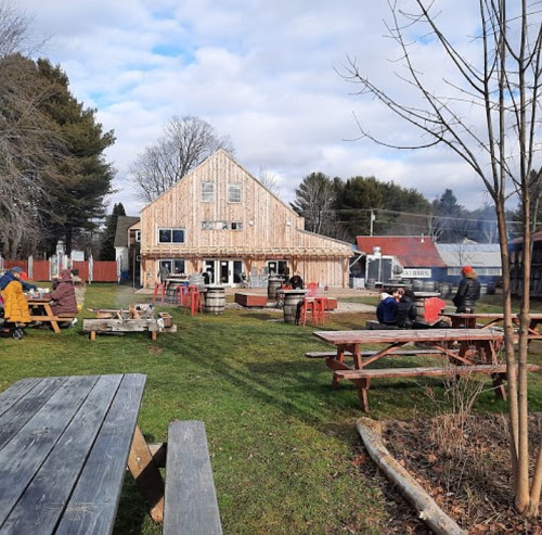 A rustic outdoor space with picnic tables, a wooden building, and people enjoying the area on a cloudy day.