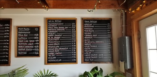A chalkboard menu displaying various bottles and four-pack options with prices, set in a cozy, rustic space.