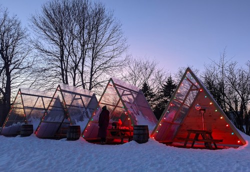 A row of triangular cabins decorated with colorful lights, surrounded by snow and trees at dusk.