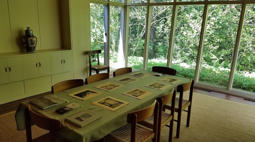A dining table with photographs laid out, surrounded by chairs, next to large windows overlooking a green landscape.