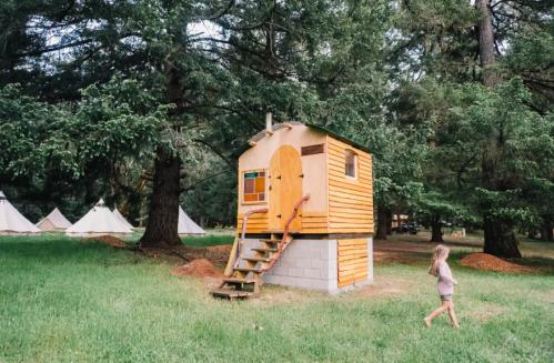 A small wooden cabin with a staircase, surrounded by trees and tents, with a child walking nearby.