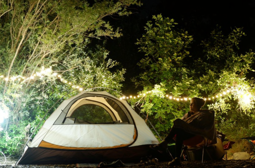 A person sits near a tent surrounded by trees, illuminated by string lights at night.