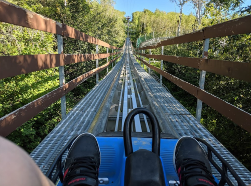 View from a ride on a mountain coaster, showing the track ahead and feet resting on the cart. Lush greenery surrounds the path.