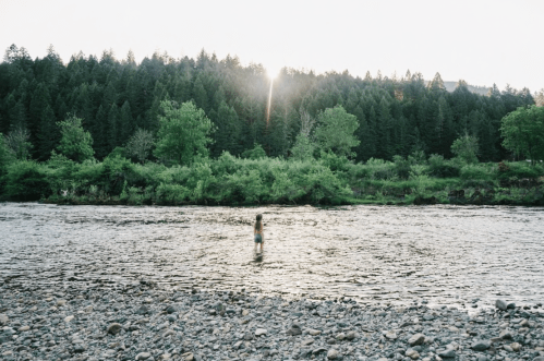 A person stands in a river surrounded by trees, with the sun setting in the background.