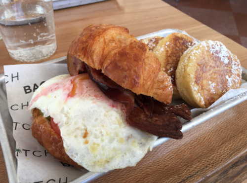 A croissant sandwich with bacon, egg, and tomato, served with two sweet pancakes dusted with powdered sugar.