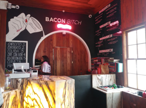 Interior of a trendy café named "Bacon Bitch," featuring a neon sign, wooden decor, and a menu on the wall.