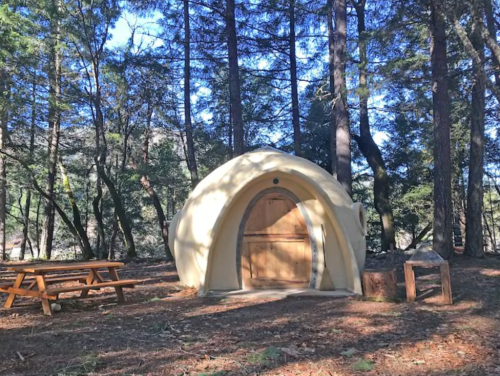 A small, rounded cabin nestled among trees, with a wooden door and picnic table nearby.