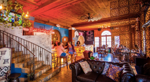 A vibrant bar interior with colorful lighting, cozy seating, and people socializing at the counter.