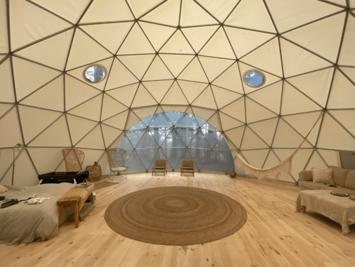 Interior of a geodesic dome with wooden floors, a round rug, seating areas, and large windows letting in natural light.