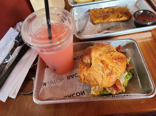 A croissant sandwich with bacon and avocado, alongside a pink drink in a clear cup, on a metal tray.