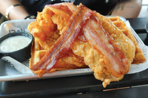 A plate of crispy fried chicken and waffles topped with strips of bacon, served with a side of dipping sauce.