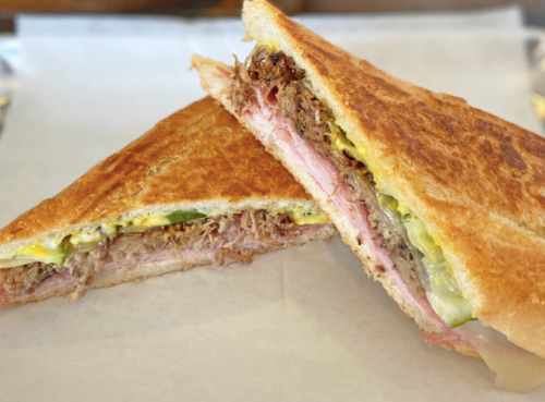 A close-up of a toasted sandwich filled with layers of meat, pickles, and mustard, cut in half on parchment paper.