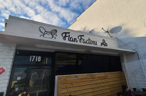Sign for "Flan Factory" on a white building with a wooden fence and blue sky in the background.
