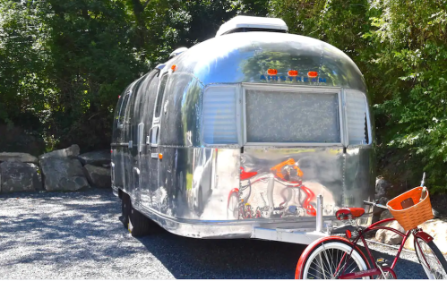 A shiny silver Airstream trailer parked on gravel, with a red bicycle and basket nearby, surrounded by greenery.