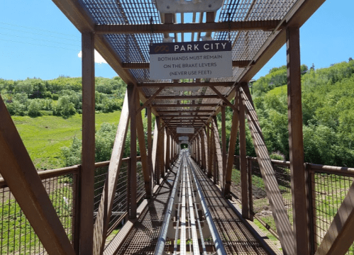 A metal bridge with signs instructing to keep hands on brake levers, surrounded by green hills and blue sky.