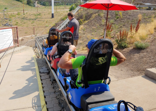 Two people sit in blue sleds at a roller coaster launch area, with a staff member nearby and a red umbrella overhead.