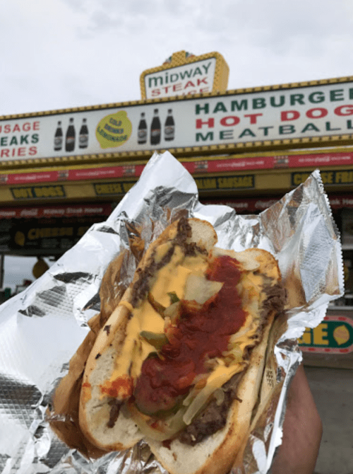 A hand holding a loaded hot dog with cheese, toppings, and sauce in front of a food stand sign.