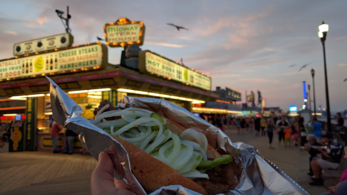 A hand holds a wrapped sandwich with onions and peppers, with a bustling boardwalk and colorful signs in the background.