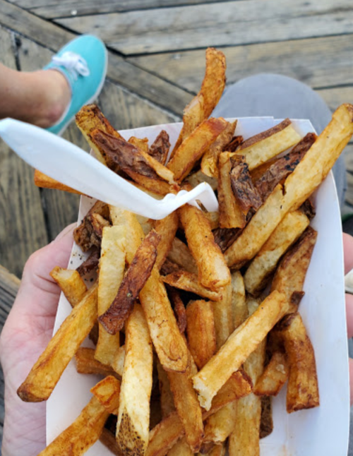 A hand holding a tray of golden-brown French fries with a plastic fork, set against a wooden surface.