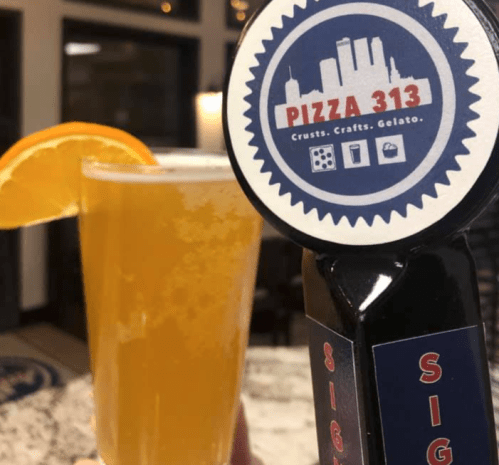 A glass of beer with an orange slice next to a tap handle featuring a logo for Pizza 313.